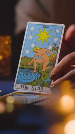 Vertical-Video-Close-Up-Of-Woman-Giving-Tarot-Card-Reading-On-Candlelit-Table-Holding-The-Star-Card